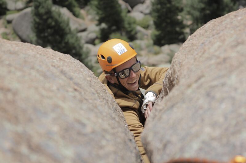 A person wearing a yellow helmet and sunglasses is wedged between two large rocks. The individual is smiling and appears to be enjoying the activity. The rocks are light brown, and the background shows trees and more rocks, suggesting an outdoor, possibly mountainous, environment. The person's clothing is a tan color, blending with the natural surroundings.